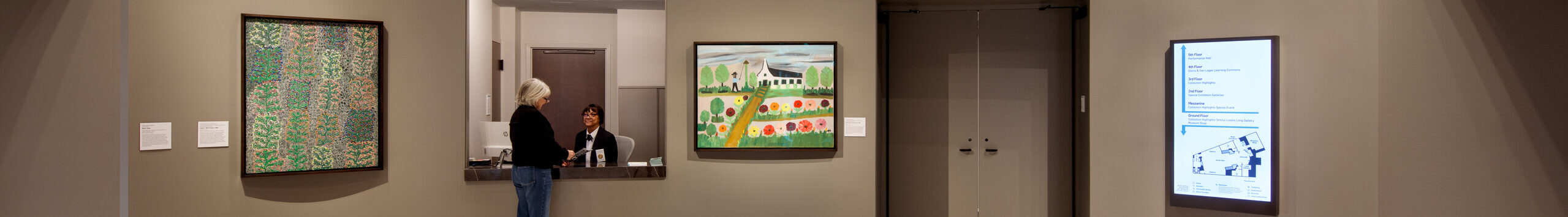 A museum rotunda features a red chandelier hanging from the center of the ceiling. The chandelier is made of red and orange glass and festooned with ornamented red-and-gold fabric garlands. LED lighting around the glass bulbs illuminates the chandelier.