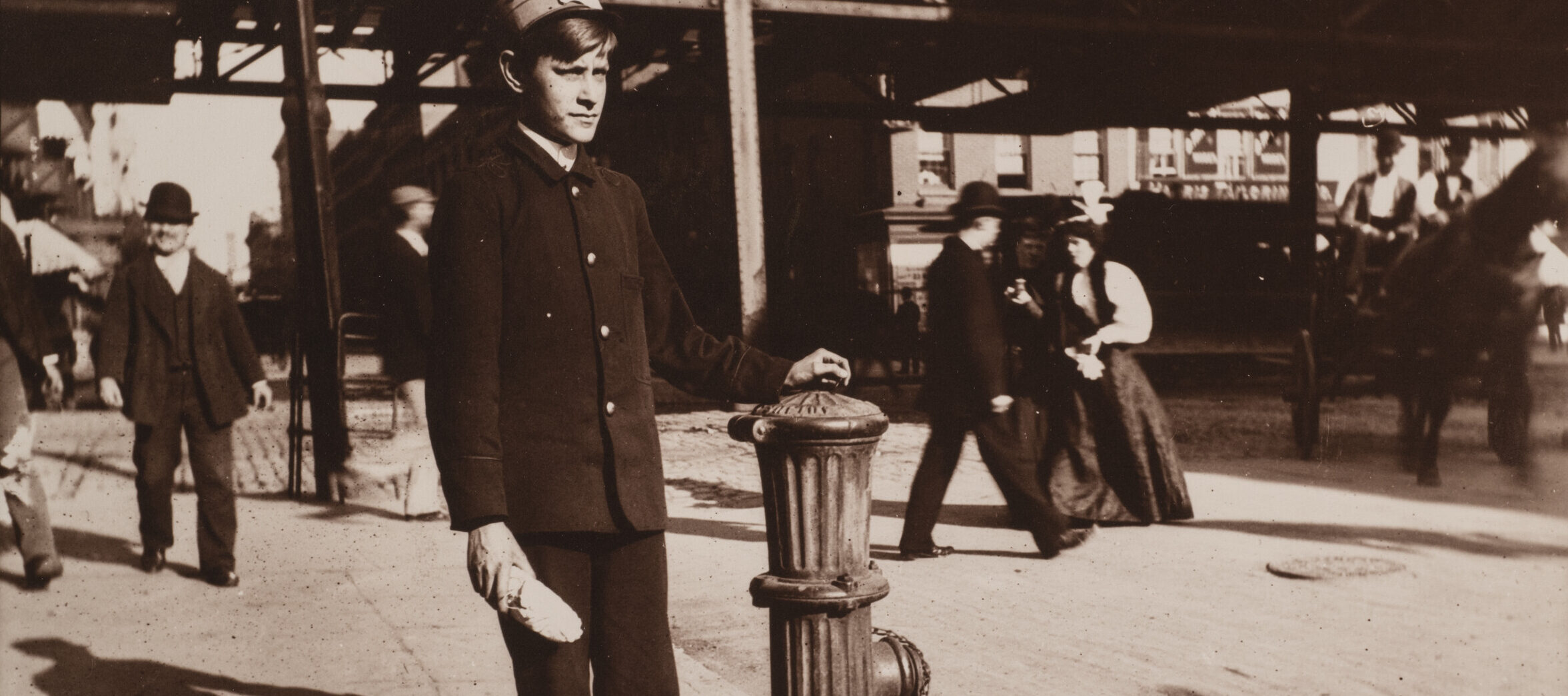 A black and white photo of a young boy in a uniform stands next to a fire hydrant on a bustling city street, holding a newspaper roll. People walk in the background, and an elevated train track is visible above.