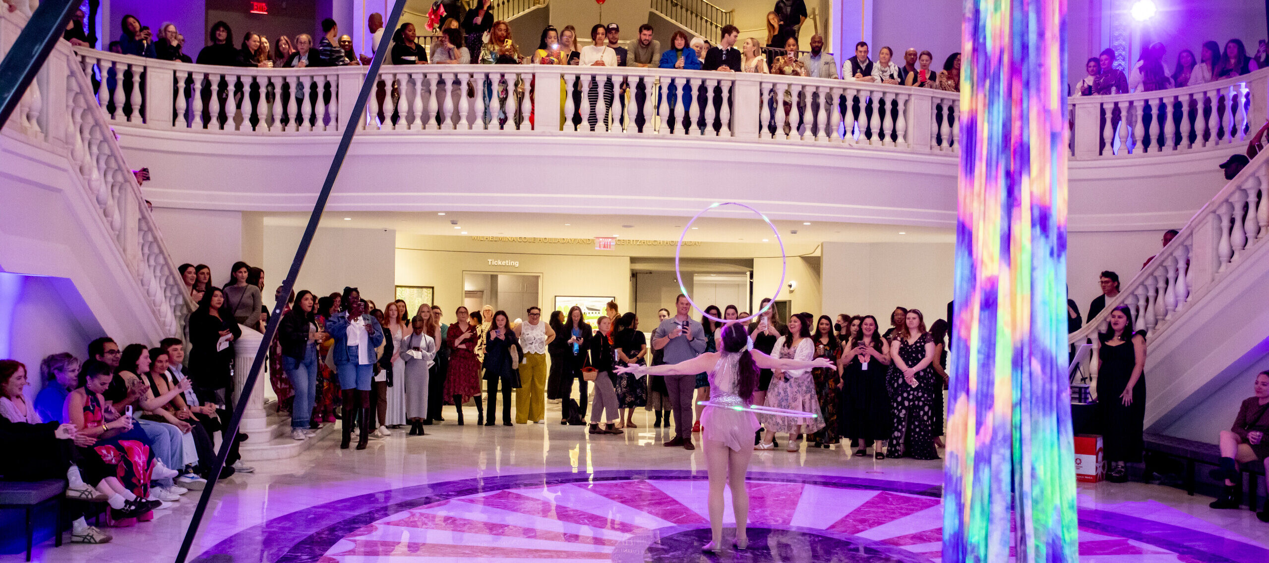 A performer with a hula hoop entertains a large crowd in an elegant hall with a marble floor, two staircases, and balconies filled with spectators under purple lighting.
