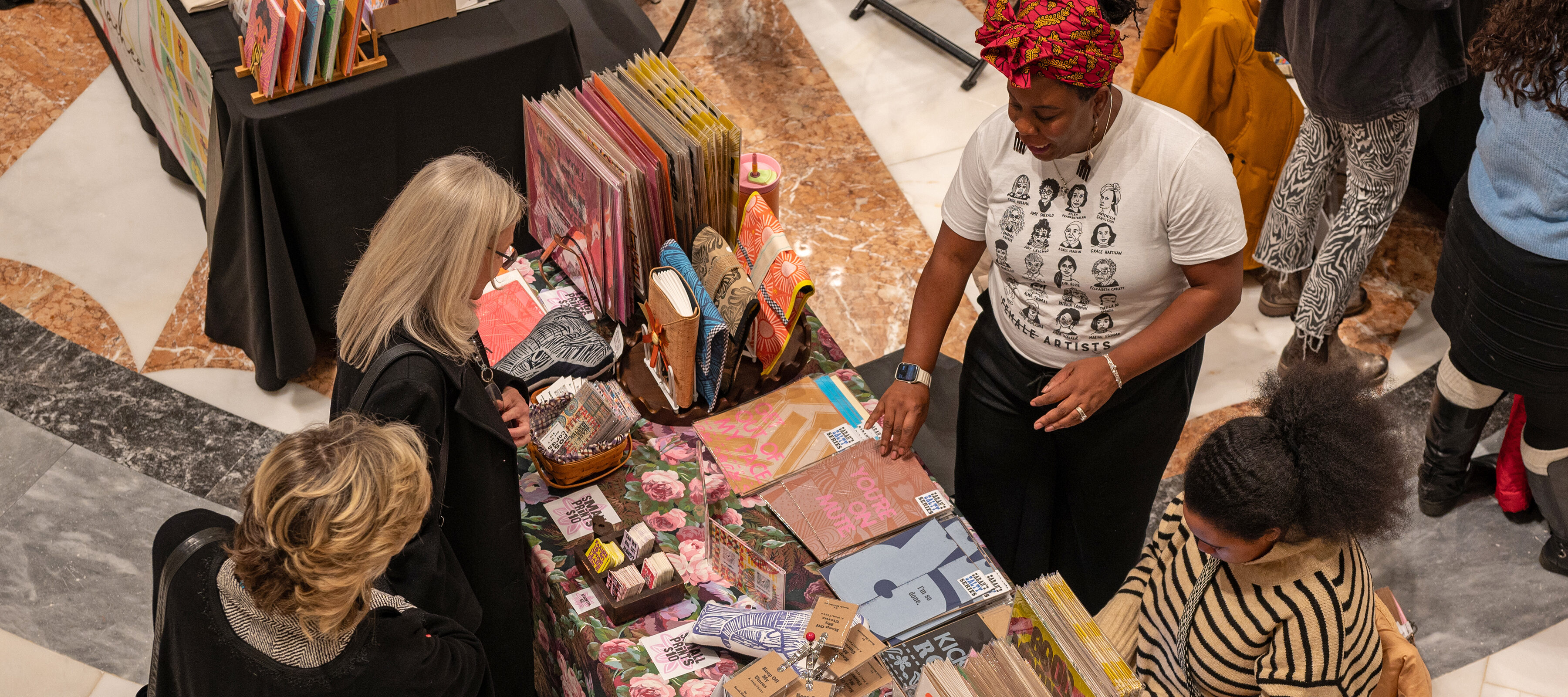 An aerial shot of two people looking at brightly colored notebooks, stickers, and prints. Two people behind the table selling the items. 