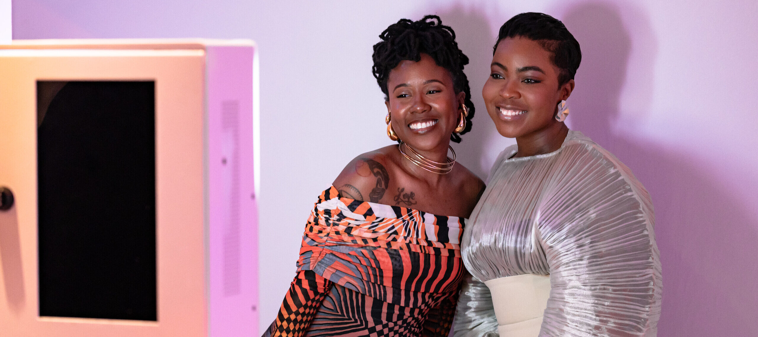 Two women with dark colored skin stand next to each other and smile in front of a white background for a photo. The woman on the left wears a gradient orange and purple dress with black stripes, and the woman on the right wears a silver and white dress.
