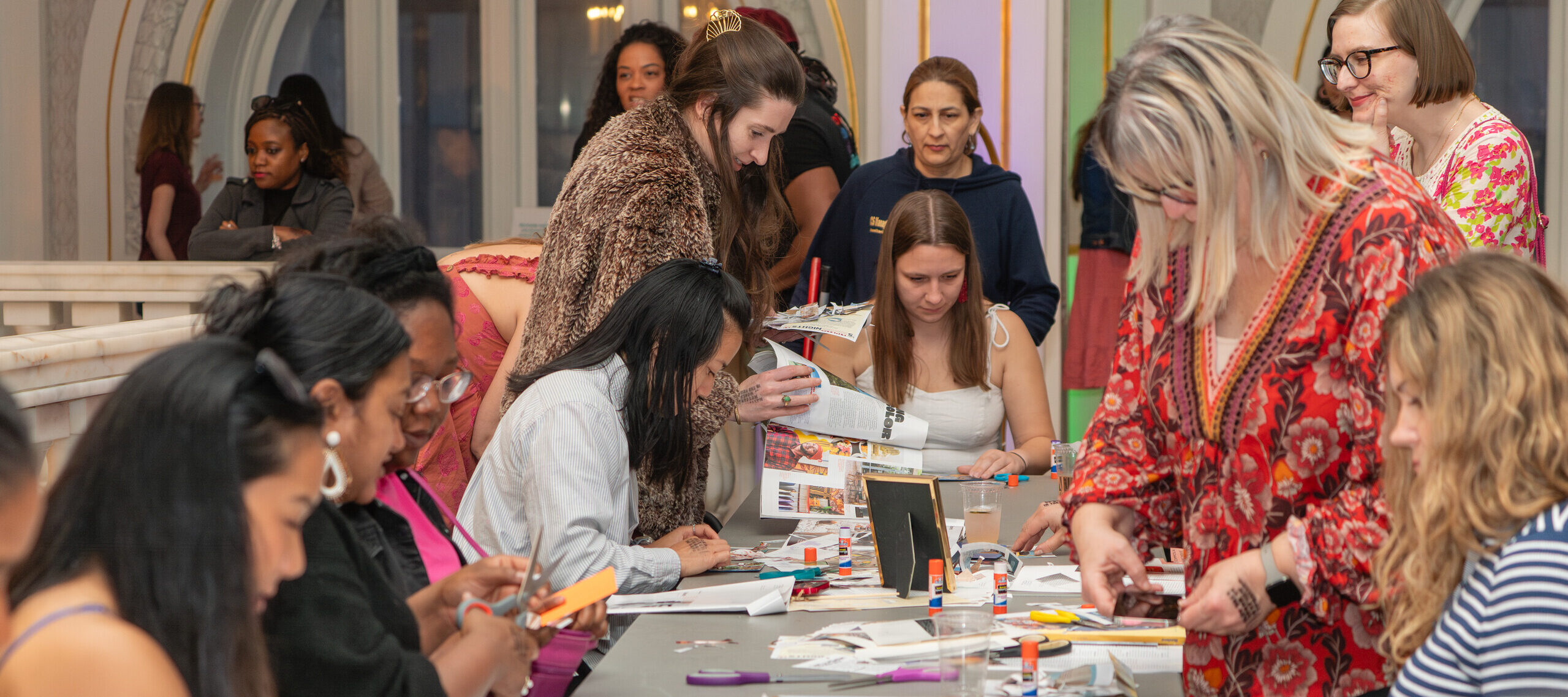 A group of women gathered around a long table work on arts and crafts projects, using magazines, scissors, and glue sticks in a bright, ornate indoor setting.