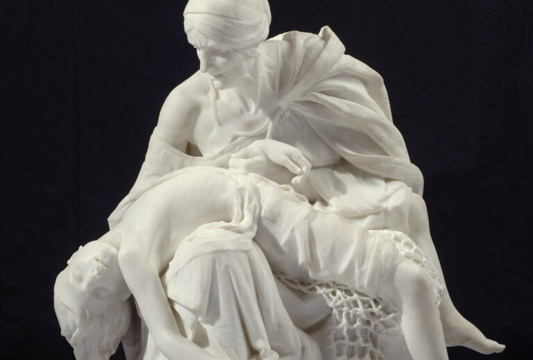 Large sculpture in white marble depicts a seated older woman cradling the body of a young boy whose limp body falls over her legs. The boy's legs are tangled in a fisherman's net while waves crash at the woman's feet.