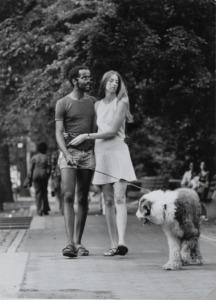 A black-and-white photo of a woman with light skin and a man with dark skin with their arms around each other while walking their dog in a park.