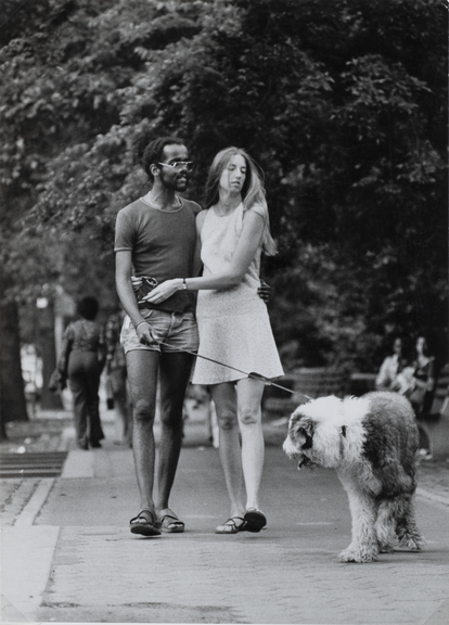 A black-and-white photo of a woman with light skin and a man with dark skin with their arms around each other while walking their dog in a park.