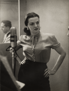 A black-and-white photo centers on a woman with light skin and dark hair. She wears a light blouse and a dark skirt and stands in front of a microphone. A man can be seen in the mirror behind the woman.