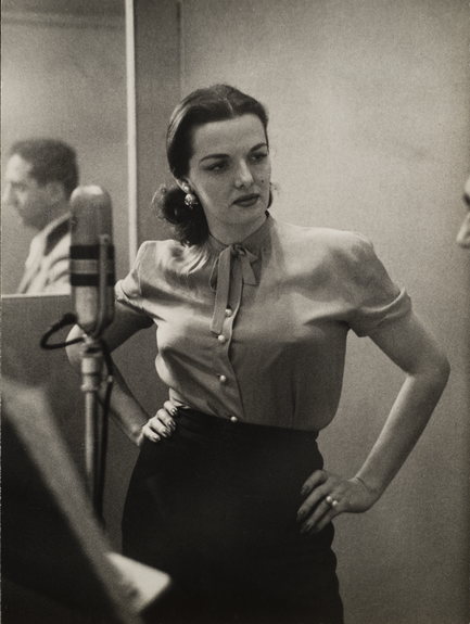 A black-and-white photo centers on a woman with light skin and dark hair. She wears a light blouse and a dark skirt and stands in front of a microphone. A man can be seen in the mirror behind the woman.