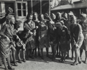 A black and white photograph of 14 women in matching uniforms, all of which have skirts covered in mud. Some women wear matching caps. Some women are touching the bottoms of their dirty skirts.