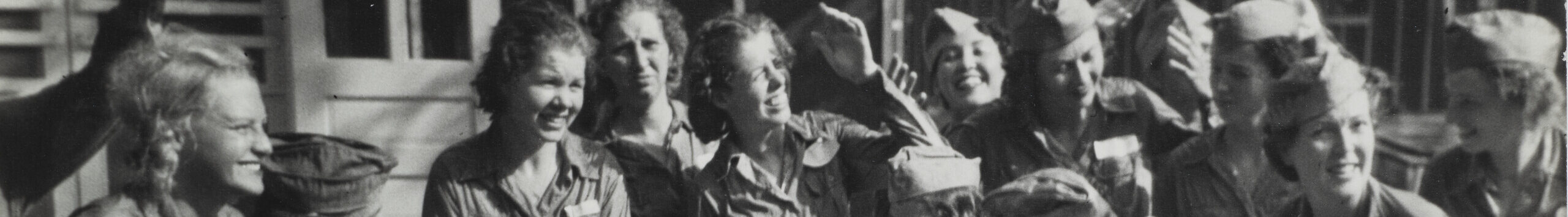 A black and white photograph of 14 women in matching uniforms, all of which have skirts covered in mud. Some women wear matching caps. Some women are touching the bottoms of their dirty skirts.