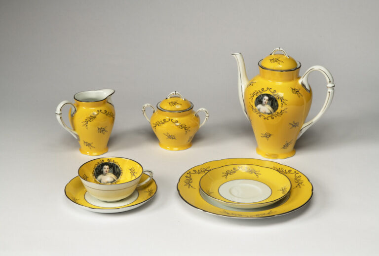 A ceramic tea set with a yellow background, gold leaf patterns, and white handles. The tea pot and tea cup feature a color image of a woman in 18th century dress.