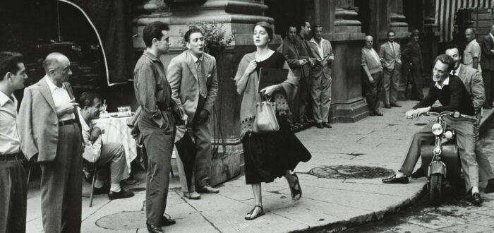 A black-and-white photograph of a young woman with light skin tone and brown hair wearing a black dress and a scarf and clutching a book and a bag walks down a city sidewalk. Several men watch her walk by, some standing against the wall of a nearby building, some sitting on chairs or motorcycles. A few men seem to be catcalling her.