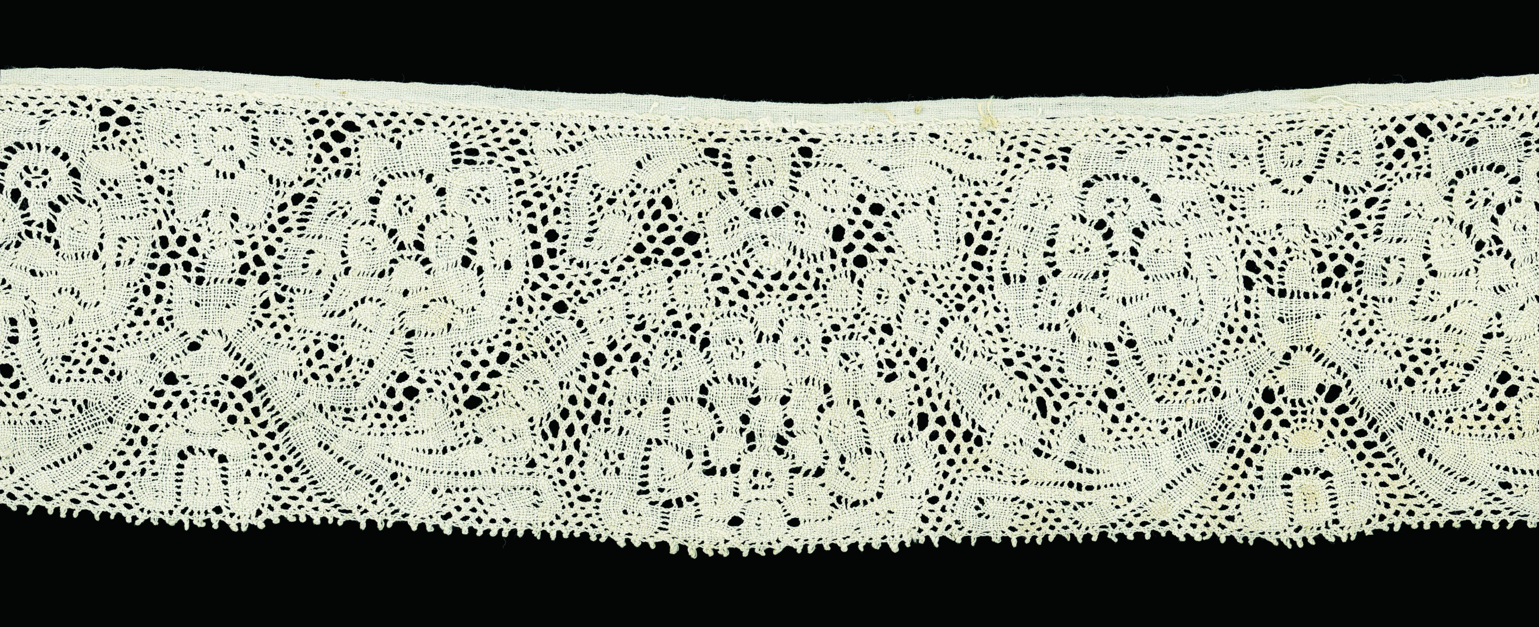 A rectangular piece of off-white lace is set against a black background. It features a closed border on one side and a fringe on the other; in between its patterns feature curling shapes and circular clusters that resemble bouquets of flowers or leafy trees.