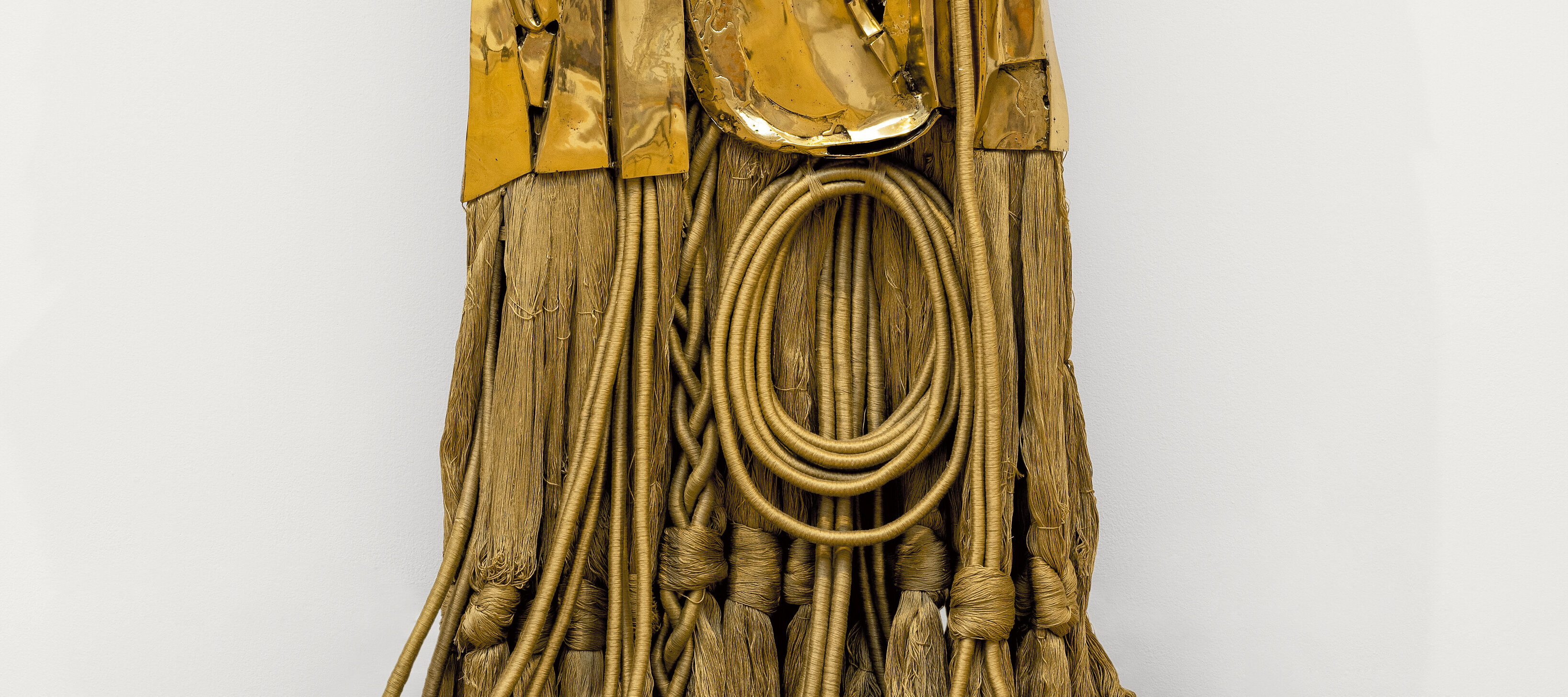 A photo of an abstract, bronze, totemic sculpture. The upper part of the sculpture looks like golden pieces of metal, while the bottom is made of folded, coiled, and braided ropes. It stands on a light wood floor against a white wall. MUST CONTACT HANNAH SHAMBROOM FOR EXTERNAL USAGE PERMISSION