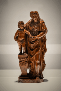 Small carved boxwood sculpture of the Virgin Mary gently holding the infant Jesus. Both figures are depicted in a tender pose, with Mary’s robes and the child’s form finely detailed in wood grain visible against a neutral background. The work is signed by the artist at the base.
