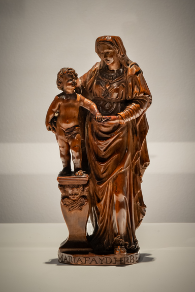 Small carved boxwood sculpture of the Virgin Mary gently holding the infant Jesus. Both figures are depicted in a tender pose, with Mary’s robes and the child’s form finely detailed in wood grain visible against a neutral background. The work is signed by the artist at the base.