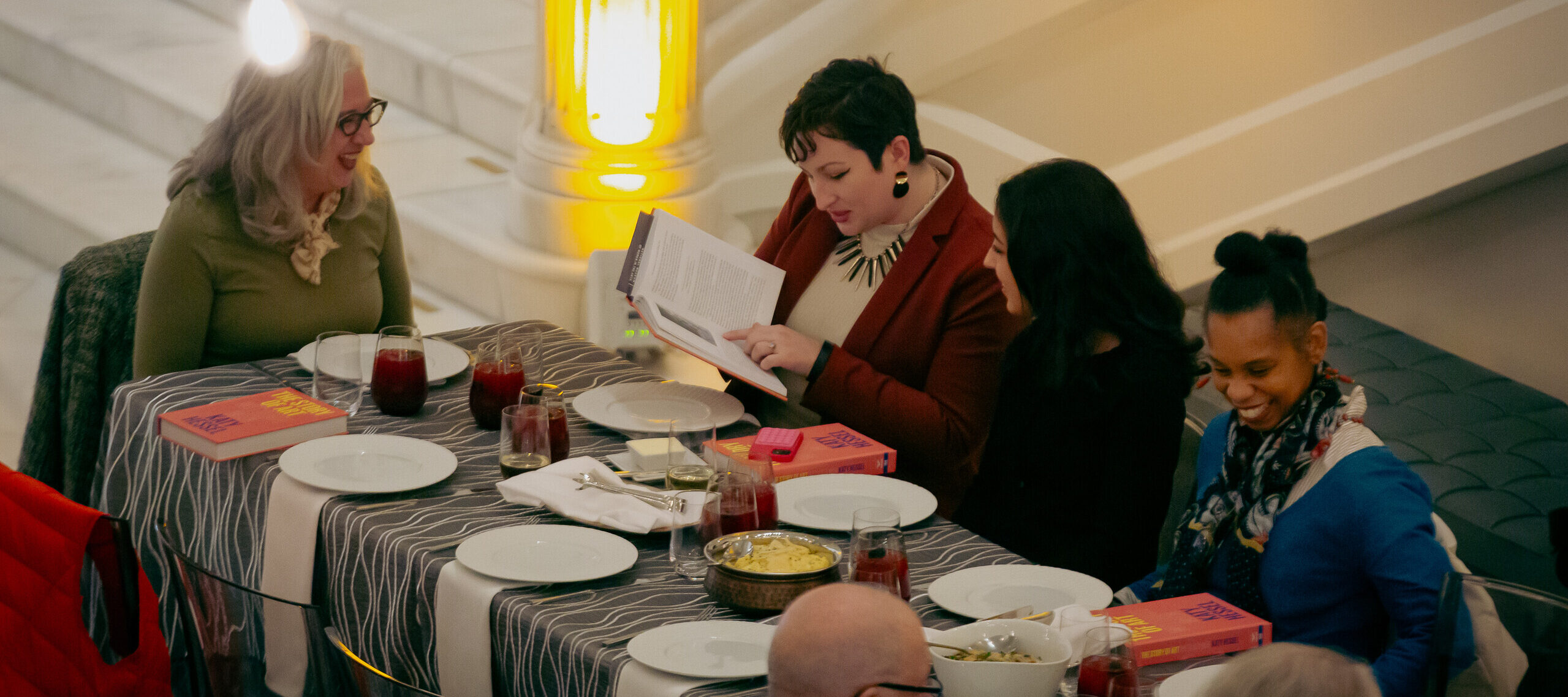 Five people are seated at a long table with a grey and white striped tablecloth, set with plates, napkins, silverware, drinks, and family-style food. A woman with short, black hair and light colored skin reads from a book.