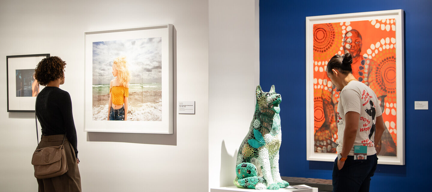Two museum visitors are walking around in a gallery. The woman to the left is viewing a photograph of a woman standing in front of a beach. The man to the right is viewing a sculpture of a dog who is wrapped up in a green and white crocheted pattern.