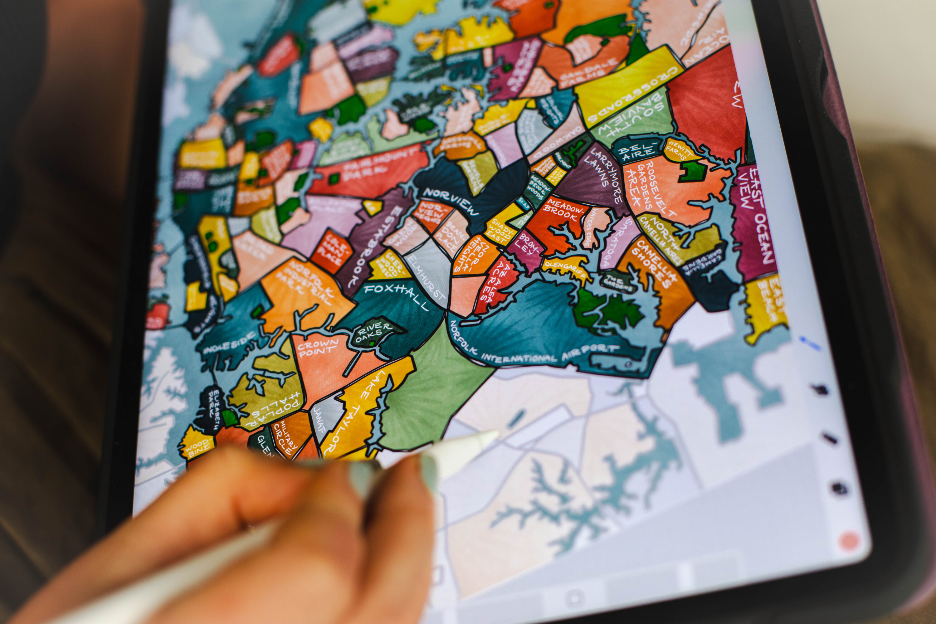A light-skinned hand holds a pen and works on a colorful map via digital drawing. The map is split into neighborhoods and each is named in white handwriting.