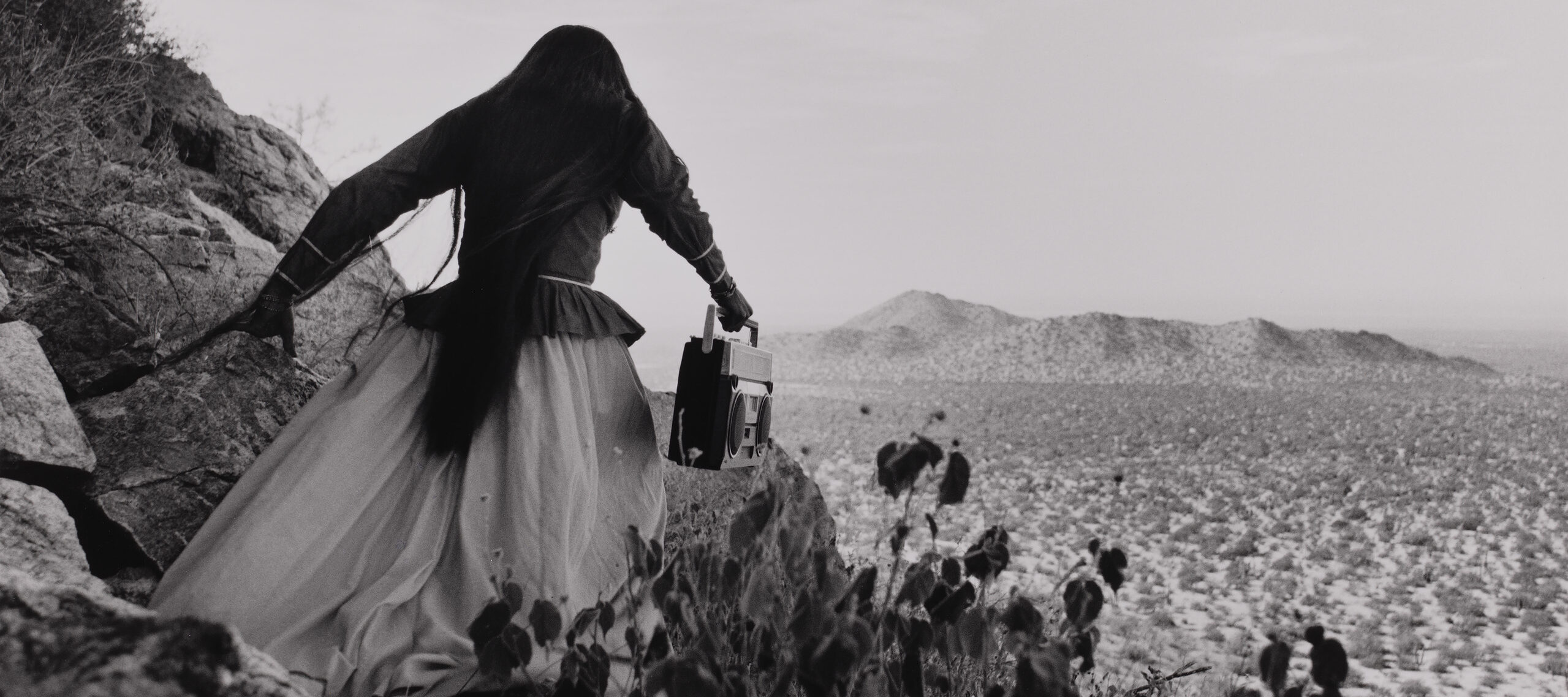 A black-and-white photograph shows the back of a woman as she crests a rocky path above a vast desert landscape beneath an expansive sky. Her traditional, ethnic full skirt, long-sleeved blouse, and long, straight, dark hair contrasts with the modern portable stereo she carries.