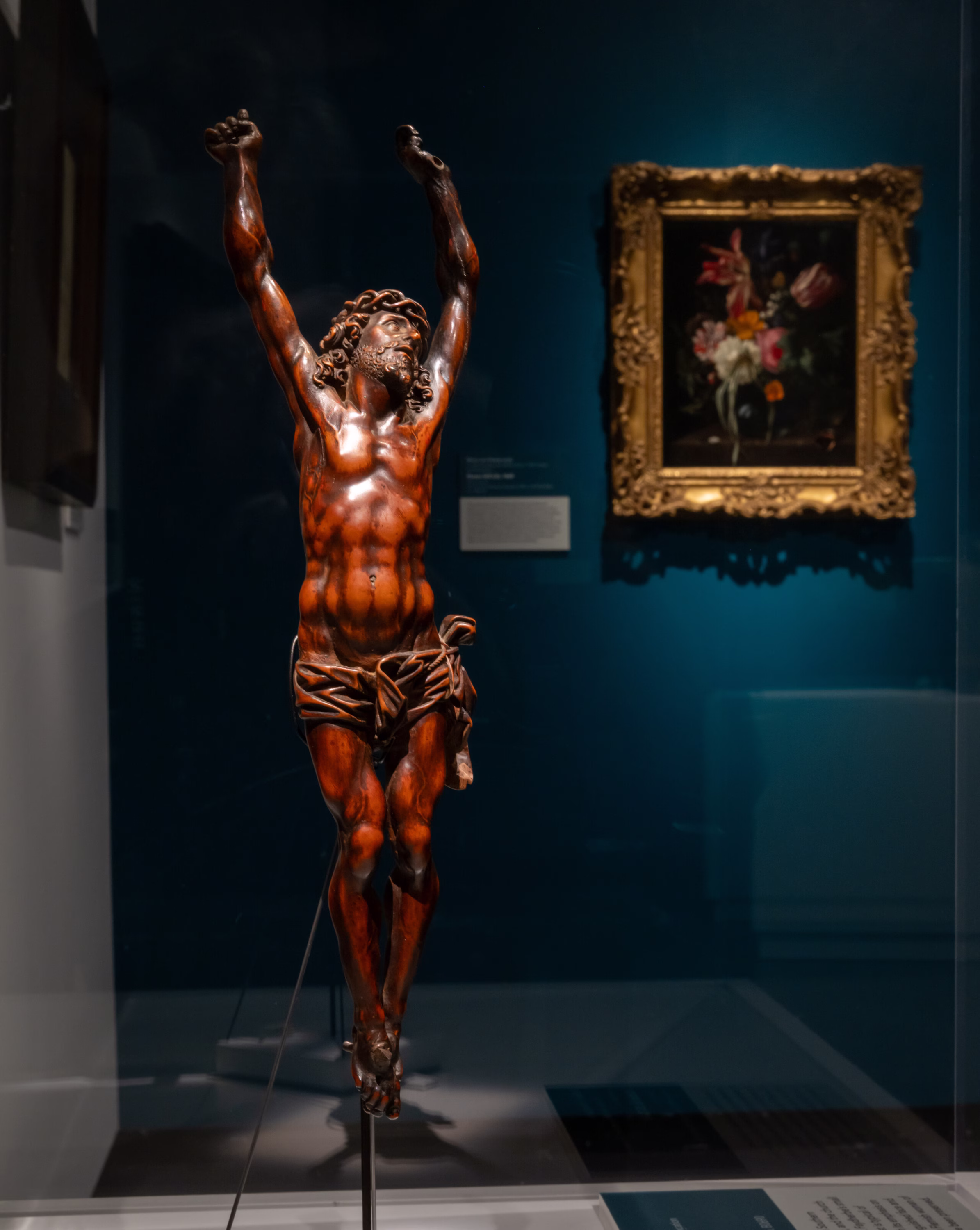A sculpture of Jesus on the cross is displayed in a museum, with arms raised and head tilted back. In the background, there is a framed floral painting on a blue wall.
