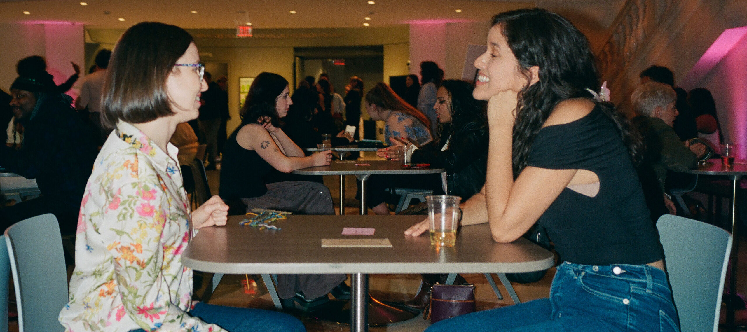 Two women sit across from each other at a small table in a brightly lit room filled with people. One wears a floral blouse and glasses, the other a black top and jeans, with a drink on the table. Background is busy with other seated groups.