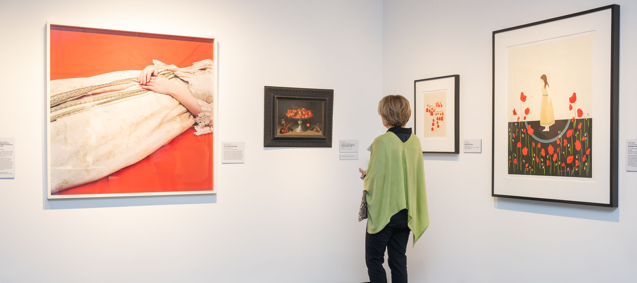 A woman with light skin tone and short light brown hair wearing a green cardigan looks at paintings in an art gallery space. One photograph is the lower half of a woman wearing a dress laying down against a orange background. A painting to the right is a bowl of cherries. The two other artworks have red flowers, one also featuring a woman standing with the flowers.