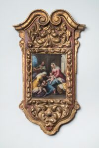 An ornate, gold-and-red wooden frame with intricate carvings surrounds a religious painting of five figures with light skin tone. The painting depicts two women seated holding babies and a man seated above observing downward.