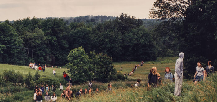 A landscape photograph featuring a field of tall green grass surrounded by leafy trees and a grey sky. A large group of people of varying ages traverse the field on foot. In the background the people form two lines, and a smaller group is congregated in the foreground.