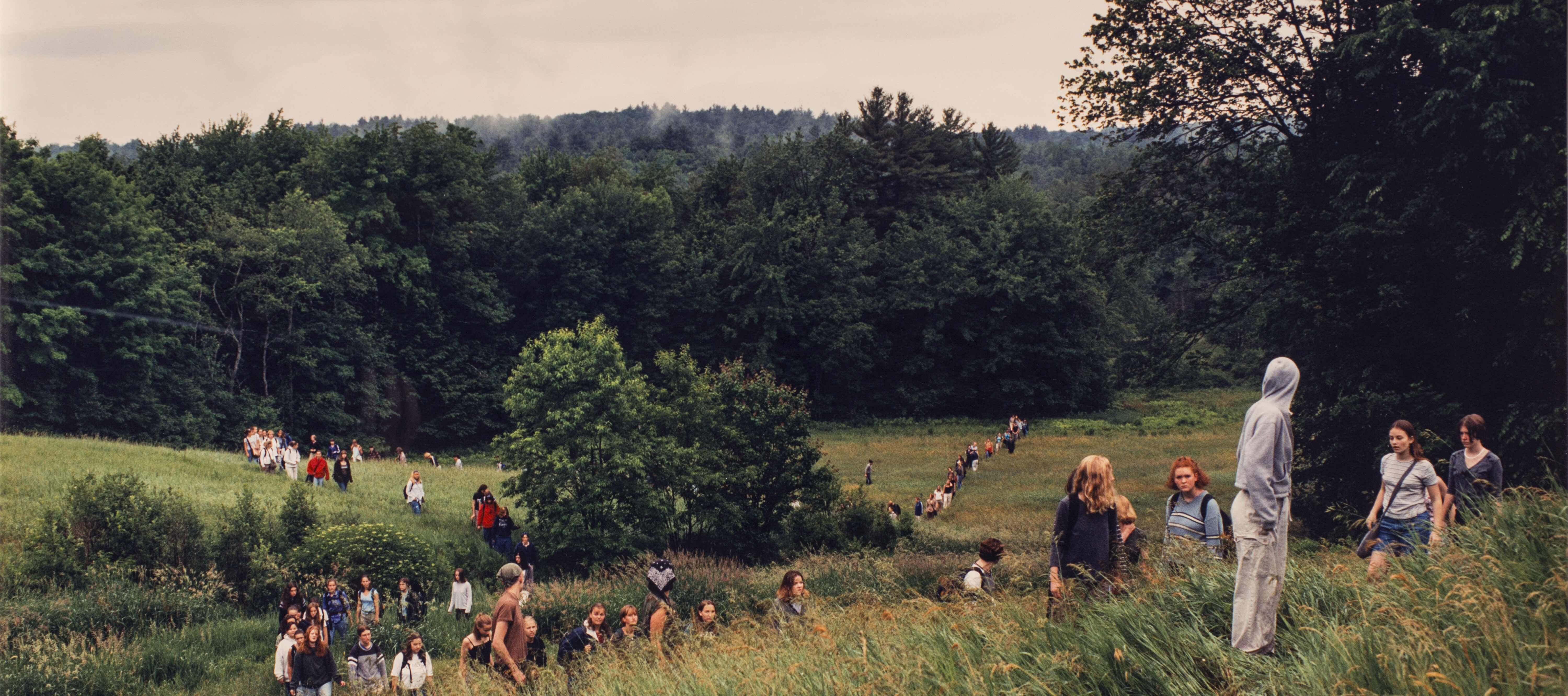 A landscape photograph featuring a field of tall green grass surrounded by leafy trees and a grey sky. A large group of people of varying ages traverse the field on foot. In the background the people form two lines, and a smaller group is congregated in the foreground.
