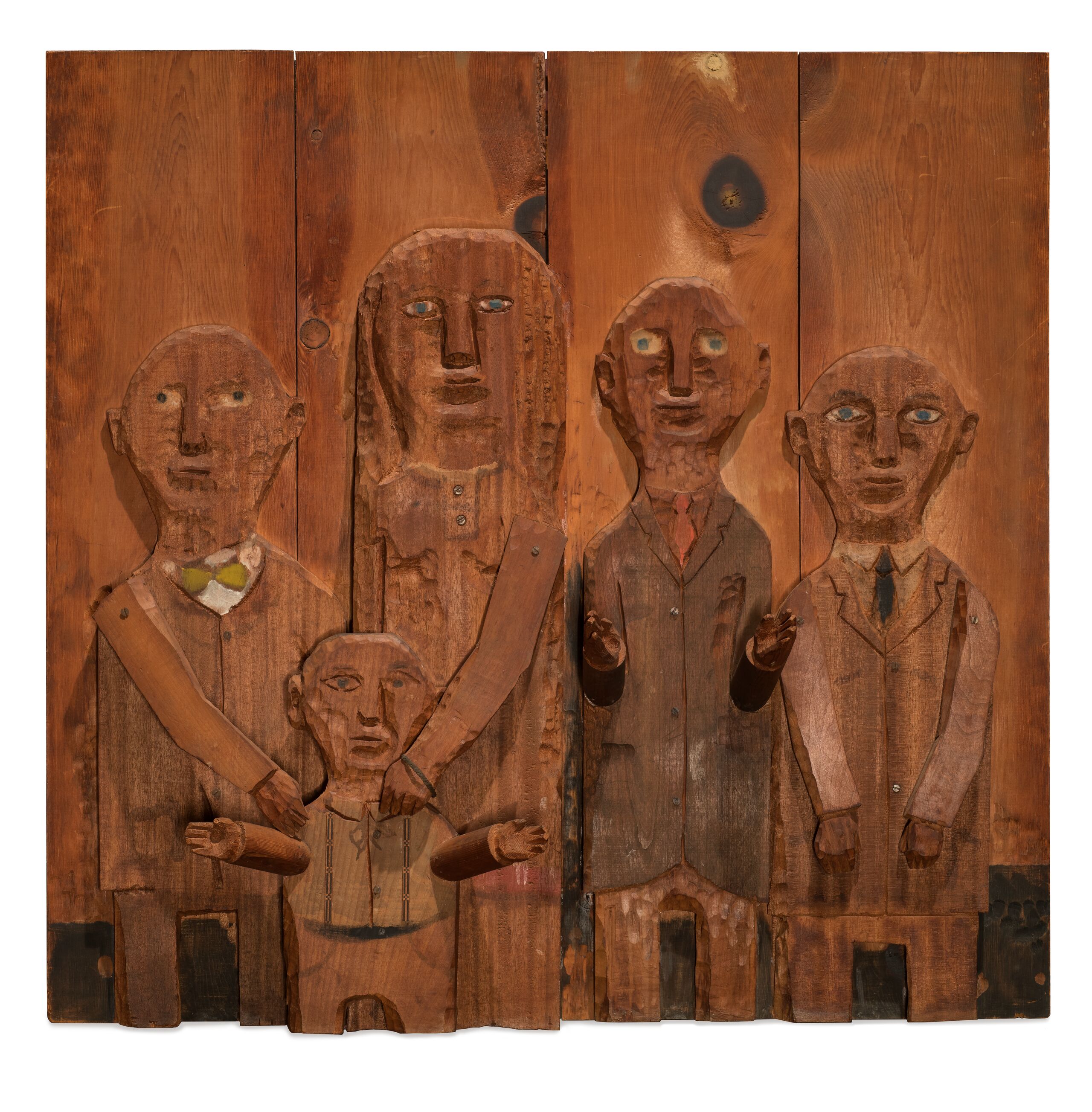 A wooden relief sculpture depicts four adults and one child. The two figures on the left put their hands on the shoulders of the child in front of them. The arms and hands of all figures are three dimensional and protrude from the work.