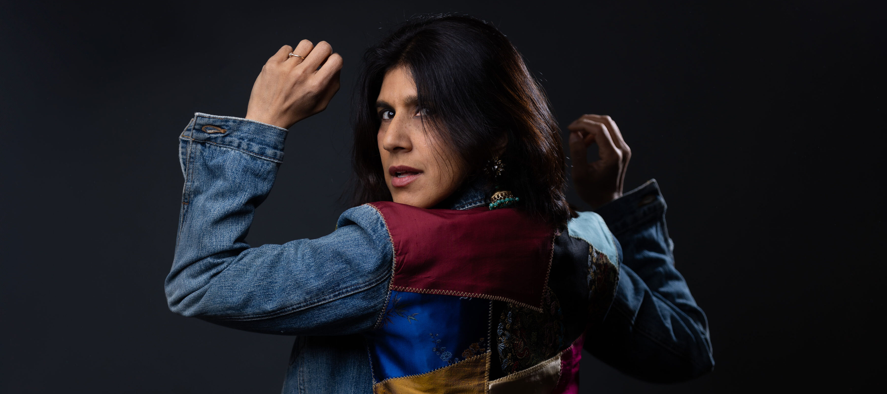 A person with shoulder-length dark hair, wearing a denim jacket with colorful fabric patches, poses against a dark background, looking back over their shoulder with one hand raised and the other near their head.