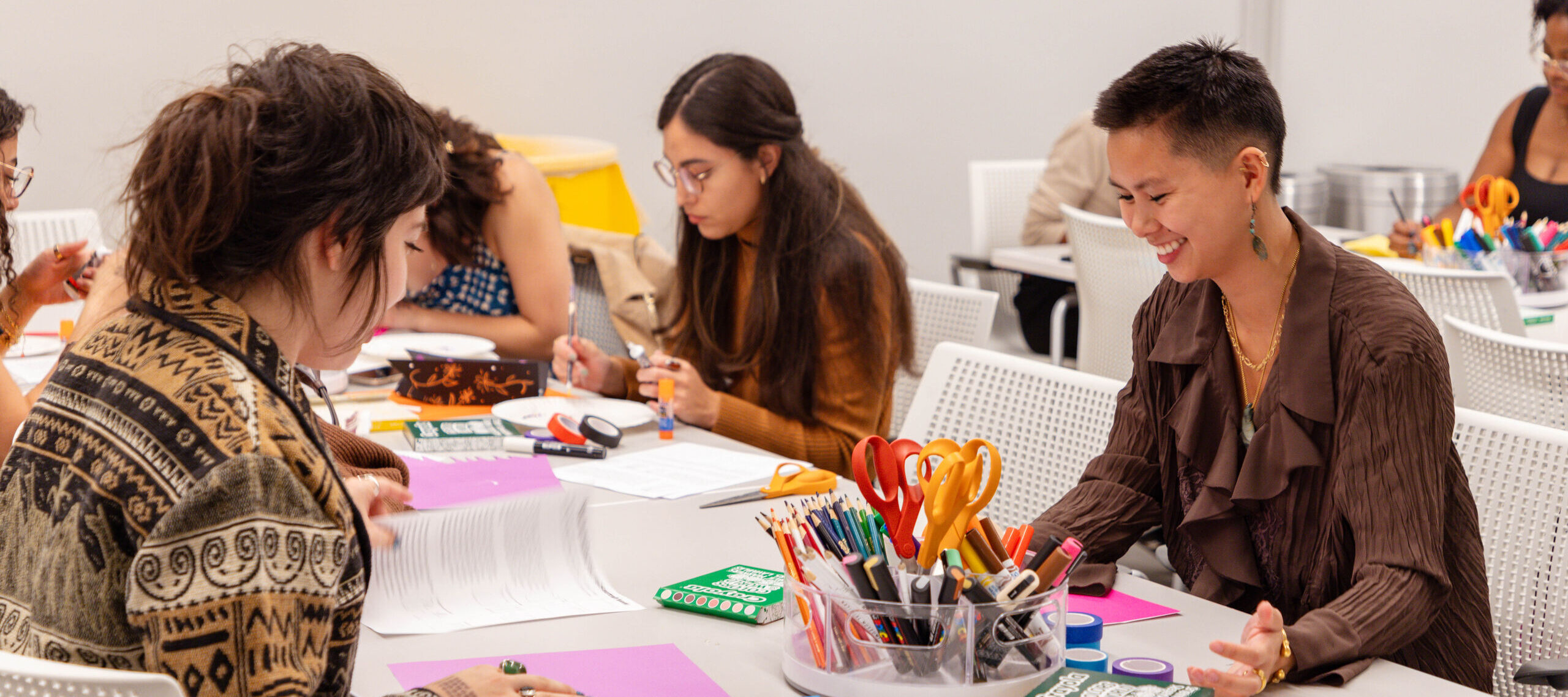 Four people sit at a table covered with art supplies, papers, and books, engaging in creative activities. One person is smiling, and the atmosphere appears collaborative and focused in a well-lit room.