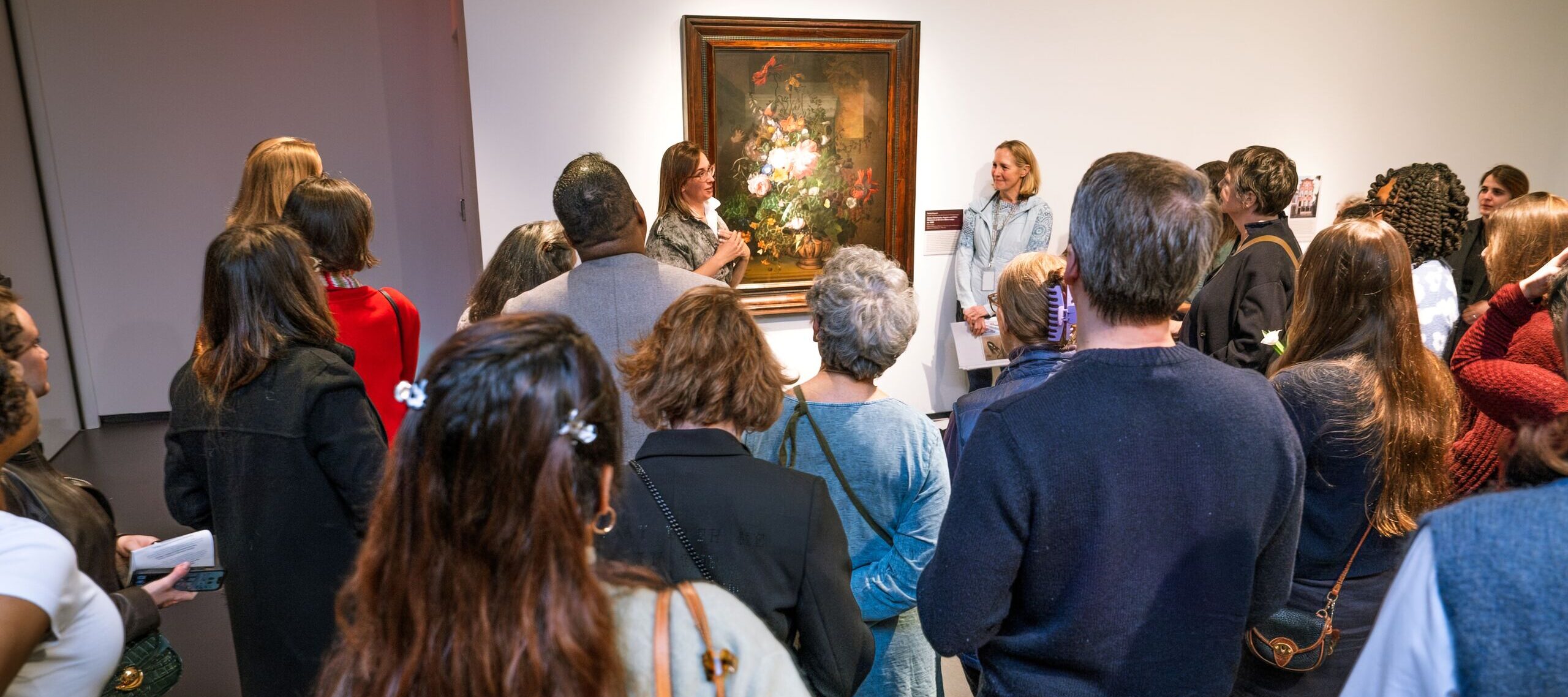 A group of people gathers around a woman giving a talk in front of a framed floral painting in an art gallery. The audience listens attentively, some holding programs or flowers.