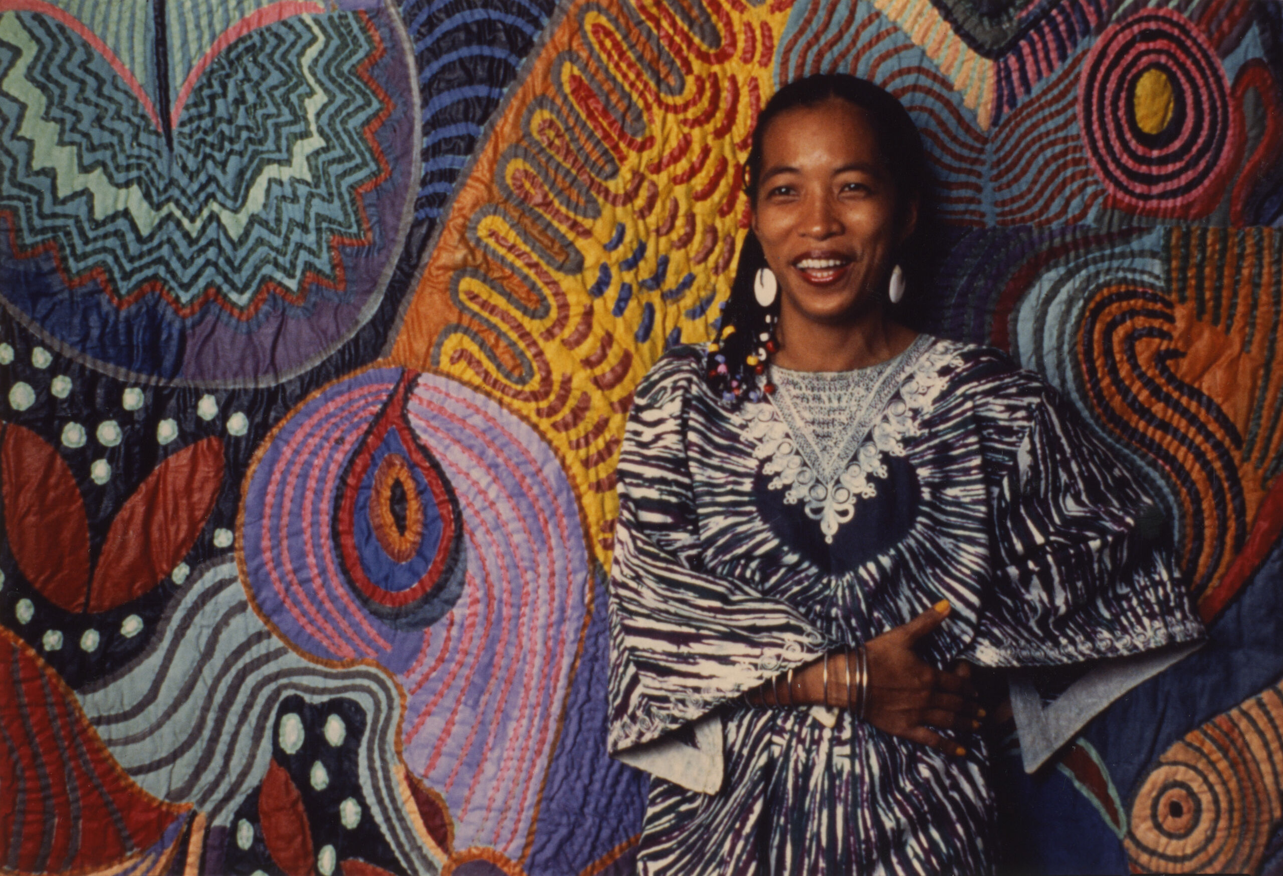 A color photograph of a woman with medium dark skin and black hair wearing a blue and white outfit and silver bangles. She stands in front of a large abstract art piece hanging on a wall.