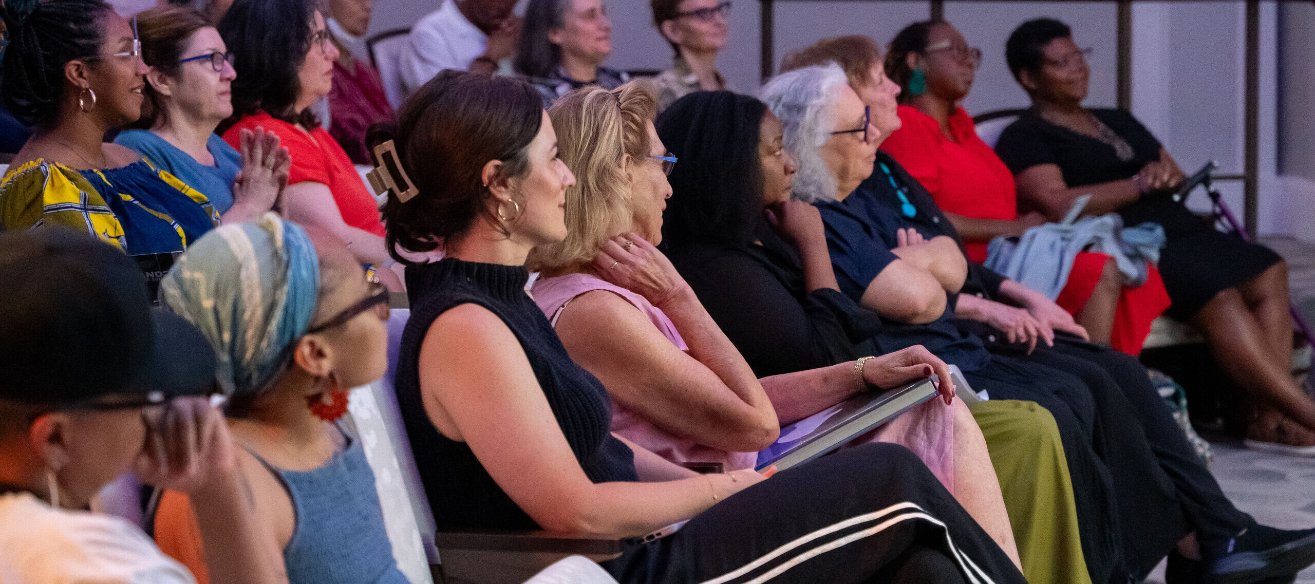A diverse group of people sit attentively in rows of chairs at an indoor event, facing forward and appearing engaged with the speaker or presentation.