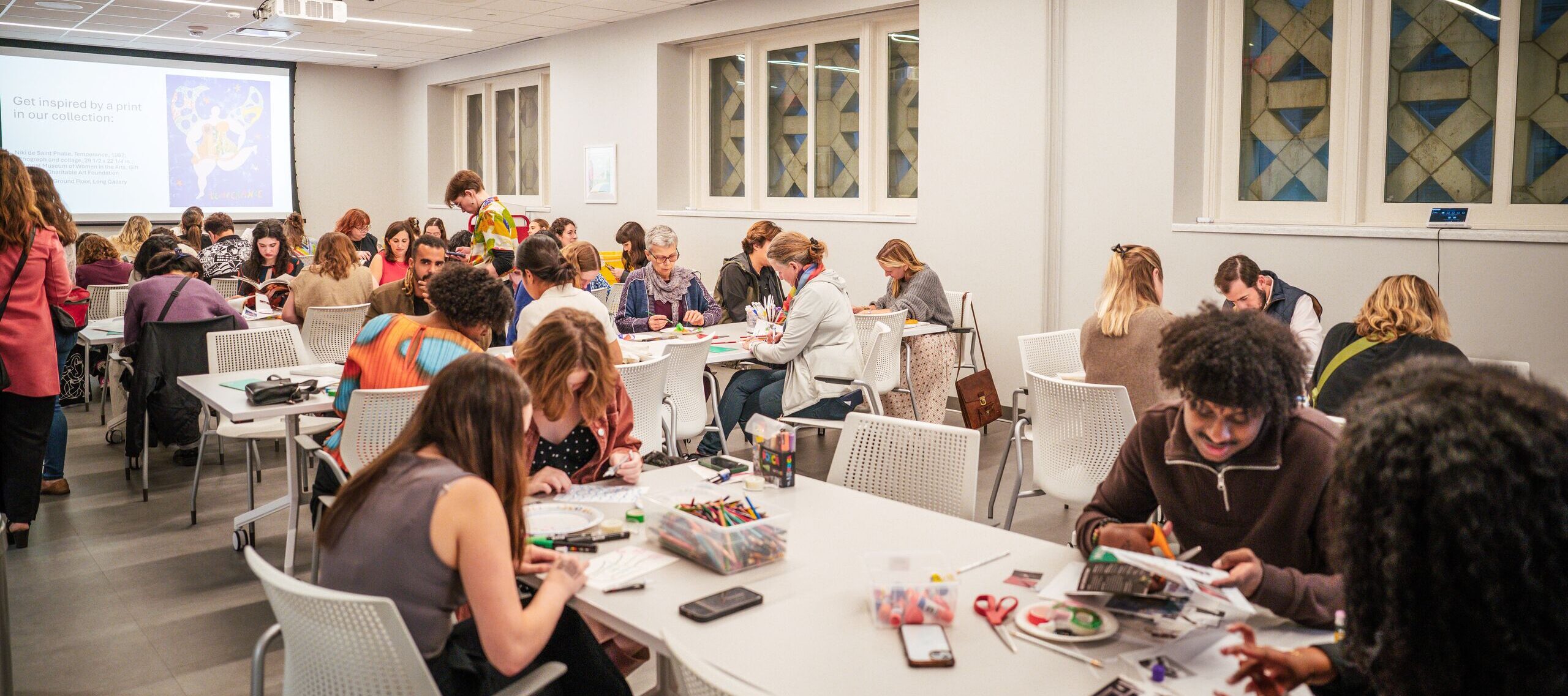 A large group of people sit at tables in a bright, modern studio classroom, working on art-making activities with colorful supplies. Others stand and talk in the background. A presentation is displayed on a screen.