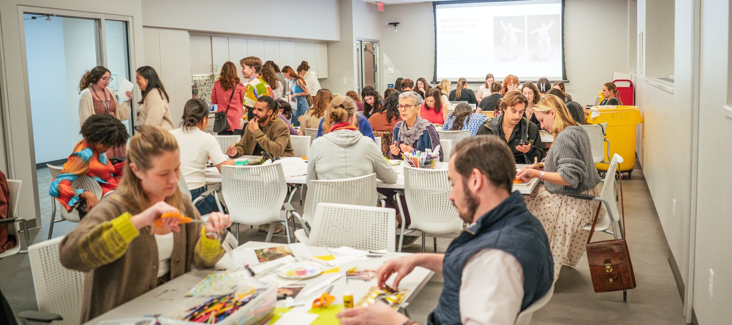 A large group of people sit at tables in a bright, modern classroom, working on arts and crafts projects with colorful supplies. Others stand and talk in the background. A presentation is displayed on a screen.