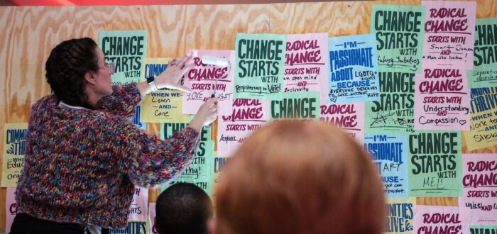 A woman pastes a paper with the text “Radical change starts with me!” onto a wall covered in colorful, handwritten posters with motivational messages, while two people look on from the foreground.