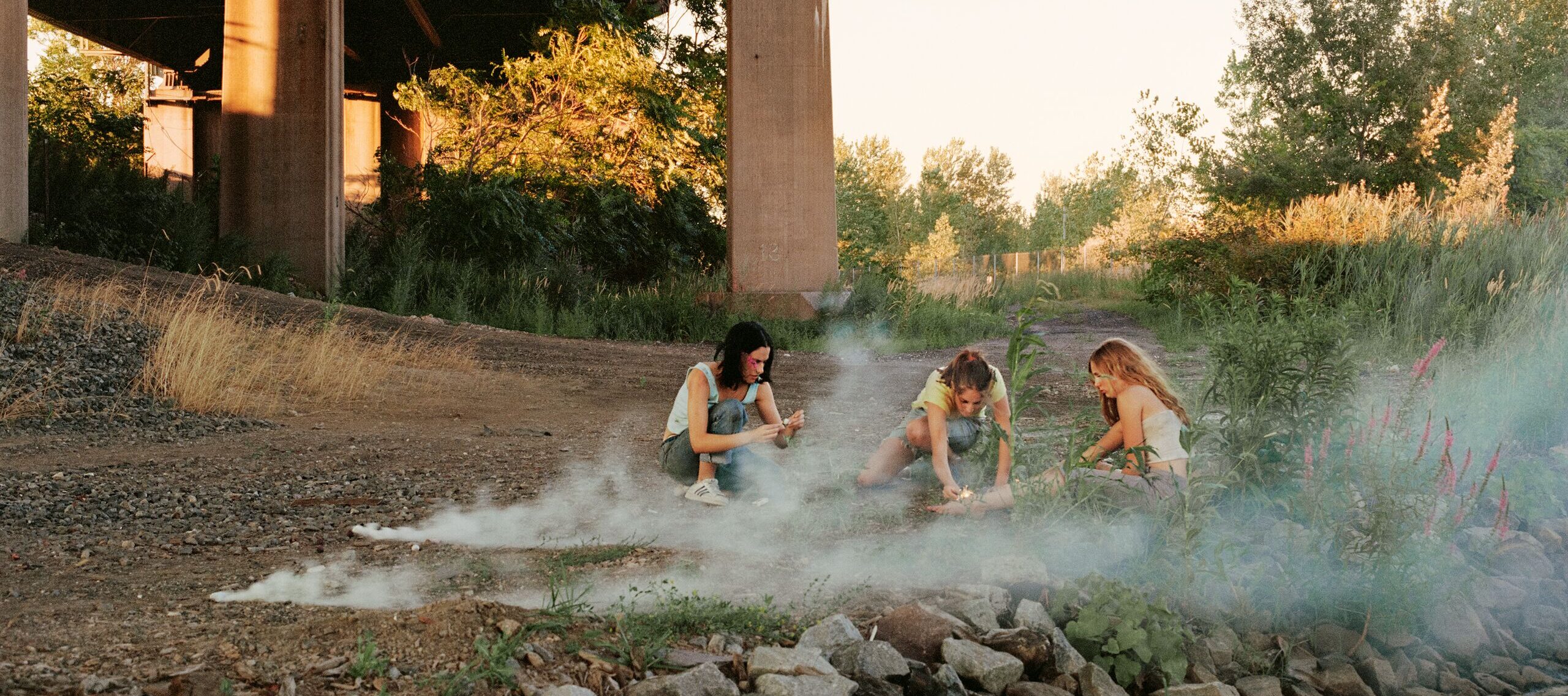 Crouching in the shadow of a highway overpass, three teenage girls with light skin focus intently on lighting smoke bombs. There are trees in the distance and wisps of white smoke surrounding them.