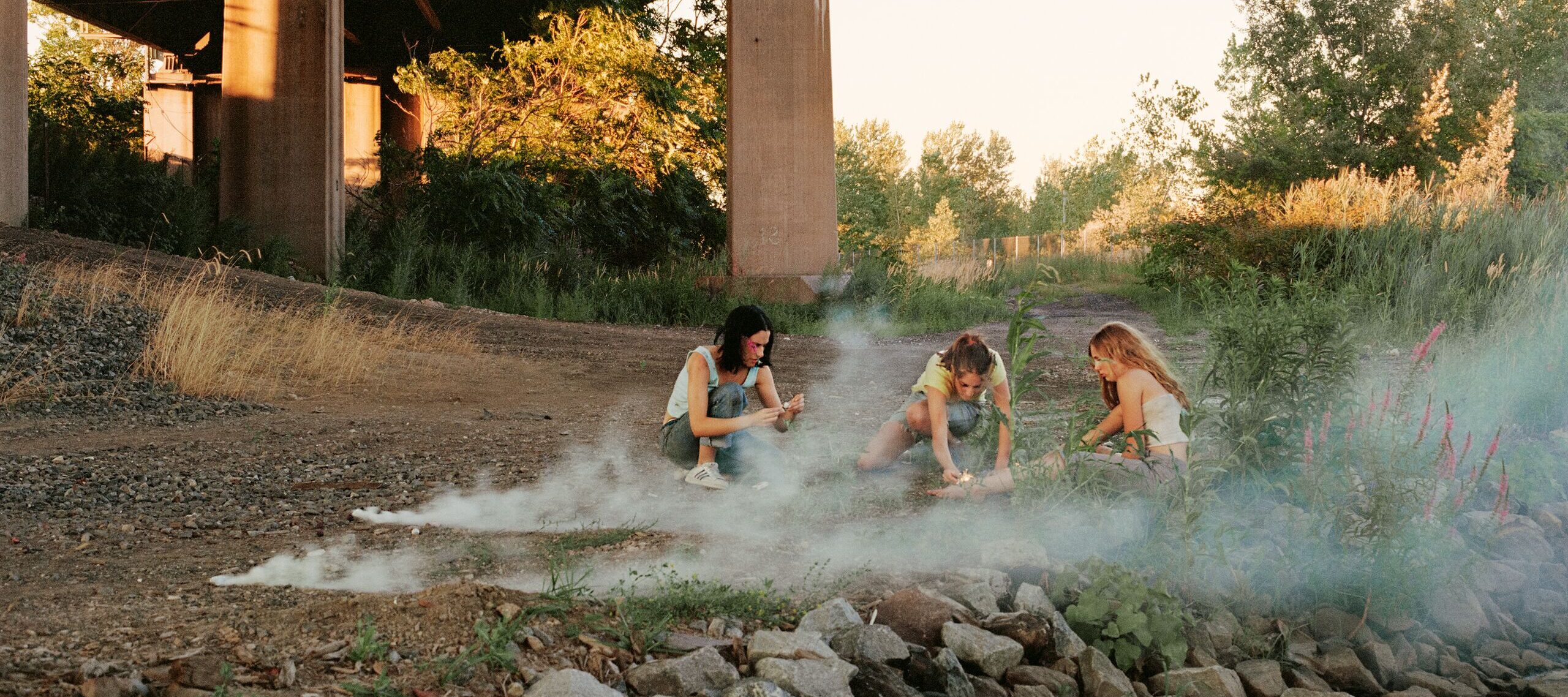 Crouching in the shadow of a highway overpass, three teenage girls with light skin focus intently on lighting smoke bombs. There are trees in the distance and wisps of white smoke surrounding them.