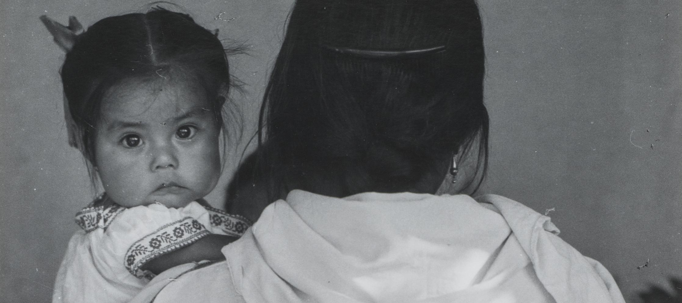 Black-and-white photograph of a woman wearing a dark skirt and light blouse holding a child. The woman faces back and her dark braid reaches past her waist. The dark-haired child stares at the camera and wears a white embroidered dress. Other figures are slightly visible beyond.