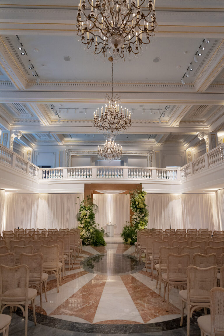 Elegant indoor wedding ceremony setup with rows of beige chairs facing a floral archway, beneath ornate chandeliers in a grand ballroom with high ceilings, marble floors, and cream-colored draped walls.
