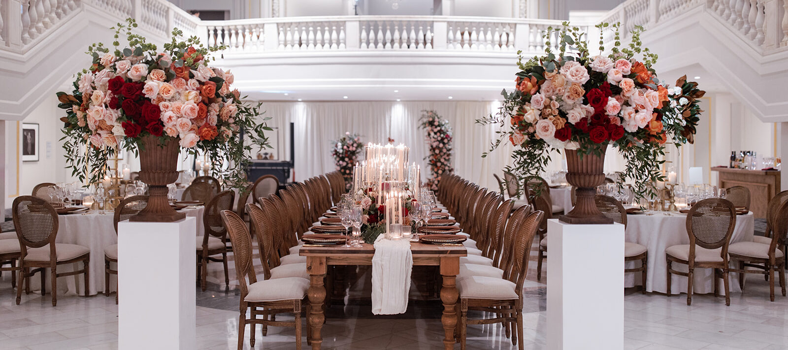 A lavish banquet hall with long wooden tables set for a formal dinner, surrounded by elegant chairs and decorated with tall floral arrangements of red, pink, and white flowers, under grand chandeliers and white balconies.