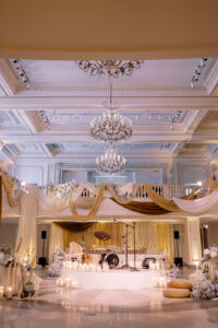 A luxurious event hall with chandeliers, draped gold and white fabric, and a stage set with musical instruments. The floor is decorated with white flowers, candles, and cream-colored cushions.