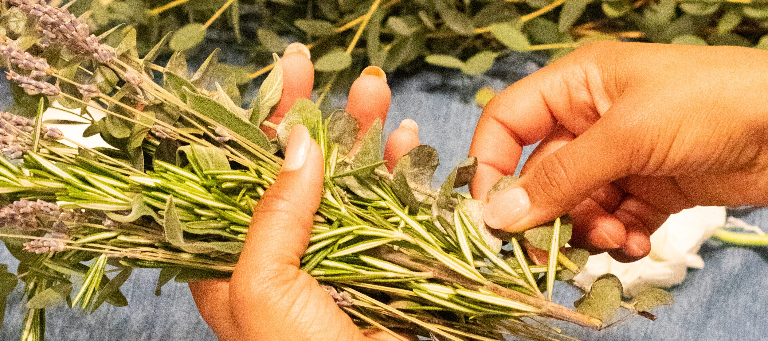 Hands assembling a herb and flower bouquet with rosemary, lavender, and eucalyptus on a blue linen surface.
