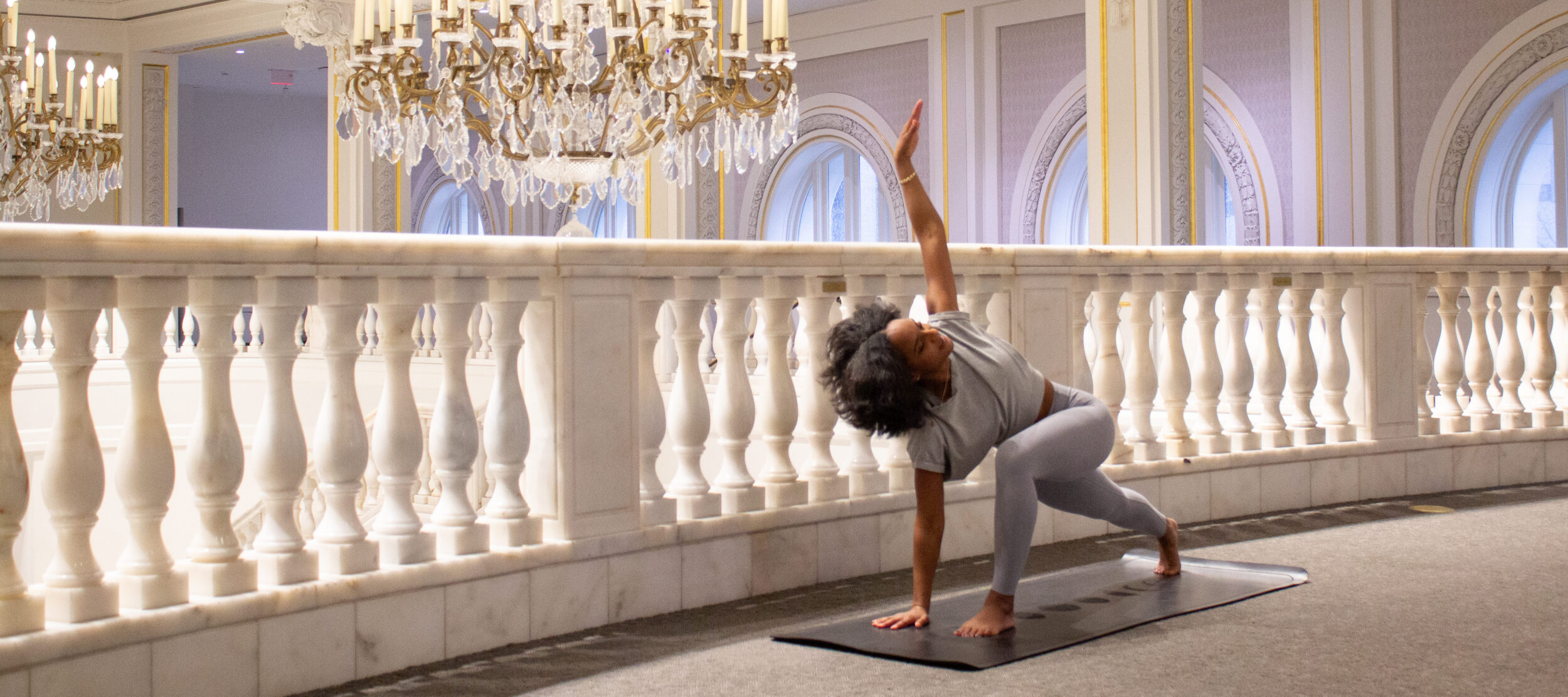 A woman practices yoga on a mat in a luxurious, ornate hall with marble railings and large chandeliers. They are in a twisting pose with one arm extended upward.
