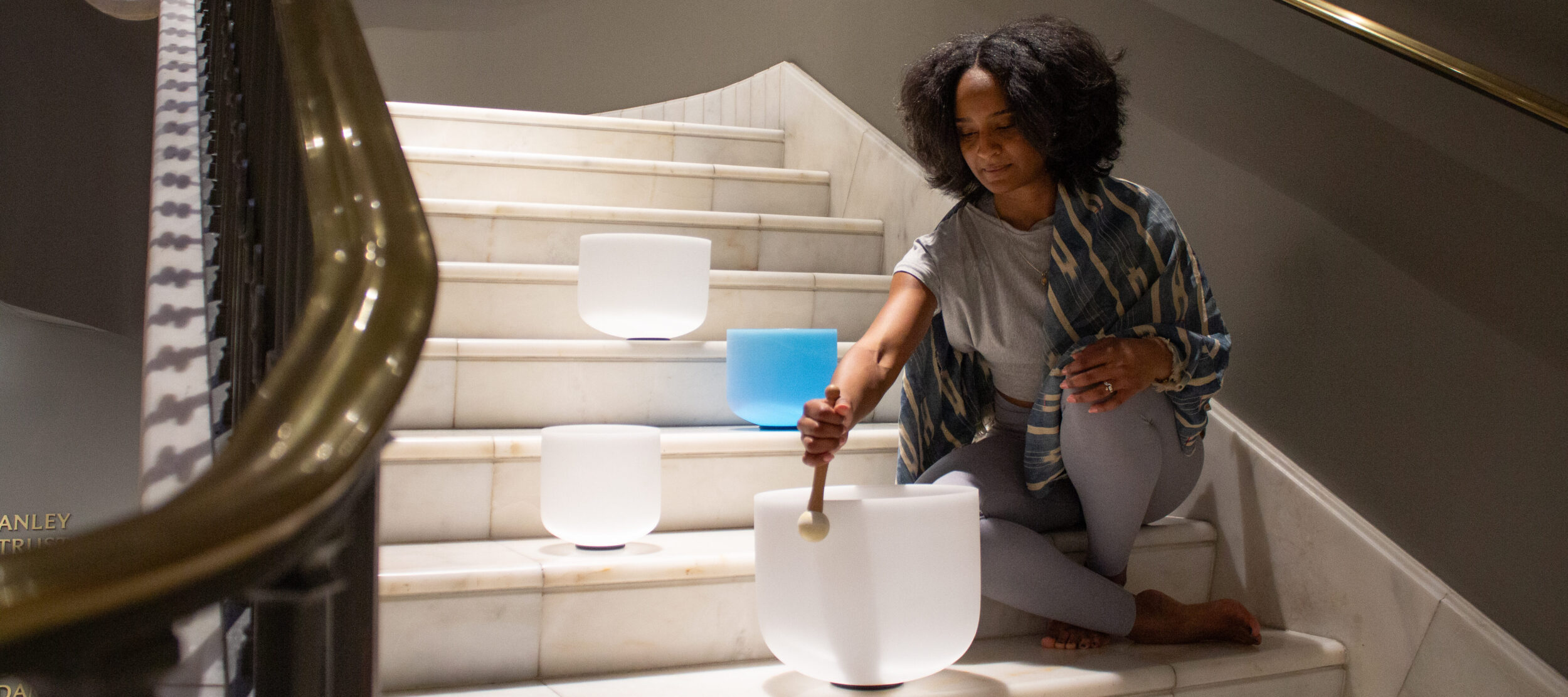 A woman sits on a marble staircase, playing crystal singing bowls with a mallet. Three bowls, two white and one blue, are placed on the steps. She is focused and appears to be meditating or performing sound therapy.