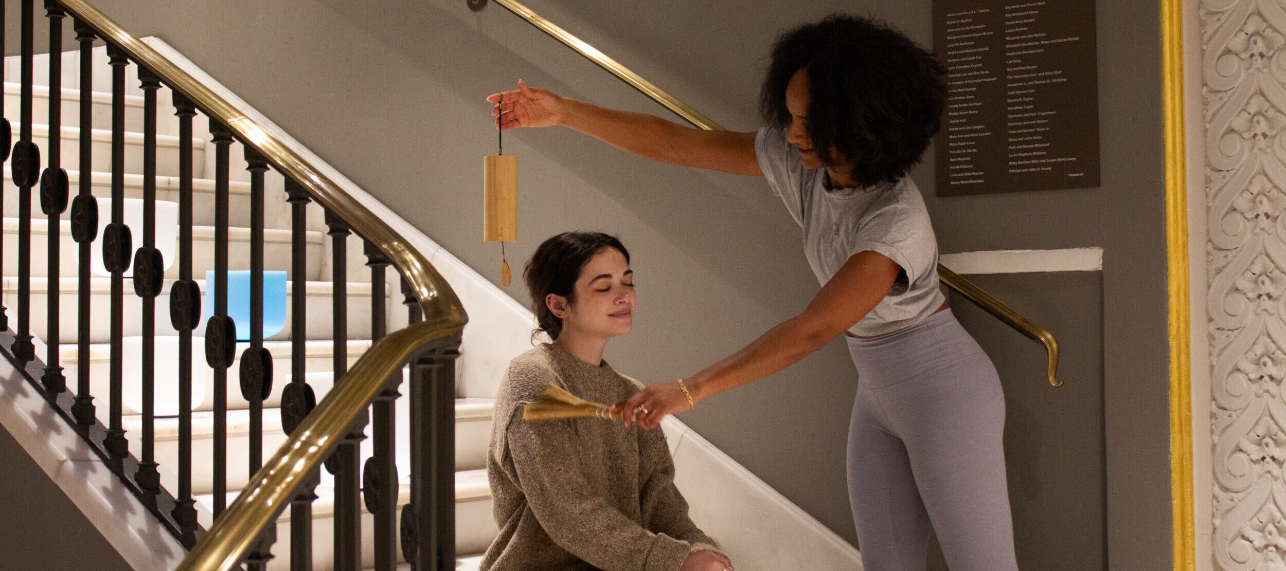 A woman sits with her eyes closed on stairs while another woman gently holds a wooden chime above her and a fan in her hand, suggesting a calming or meditative activity in an indoor setting.