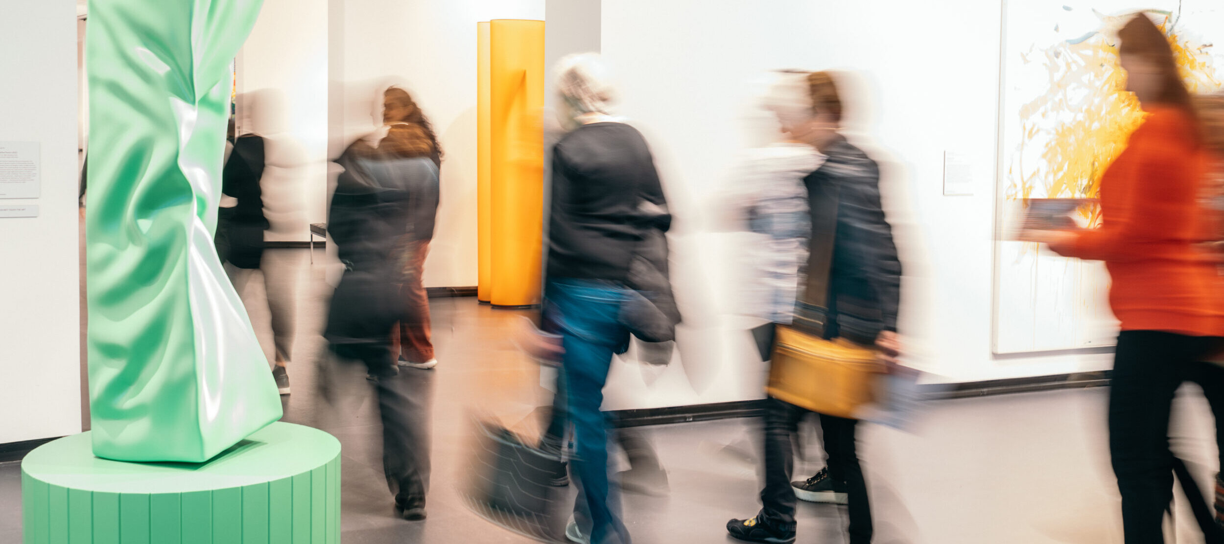 A group of museum visitors walking past a green sculpture in a modern gallery. The image has a blurred effect emphasizing the movement of the visitors from one gallery to another.