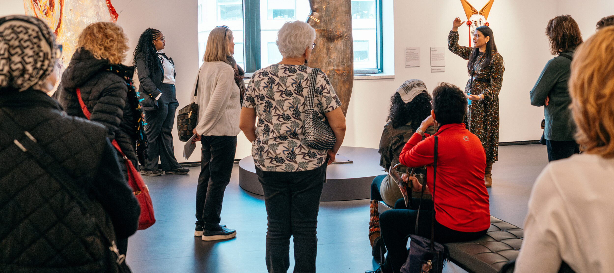 A group of museum visitors observe a large sculpture in front of a window and works of art on the wall in a modern gallery. A NMWA curator gestures next to the sculpture.